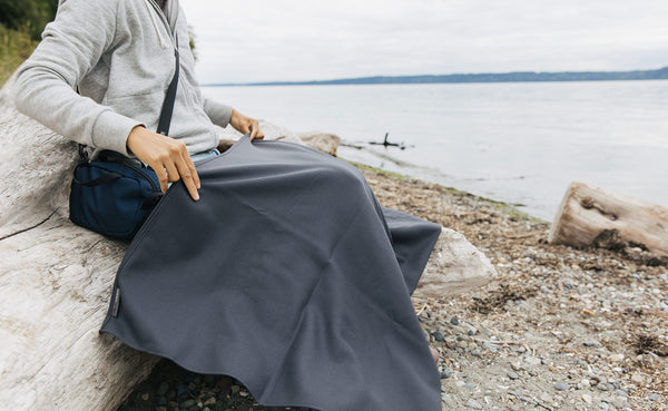 A person sitting on a log at the beach with a Large Shepherd's Wool Utility Cloth in Matter Grey on their lap.
